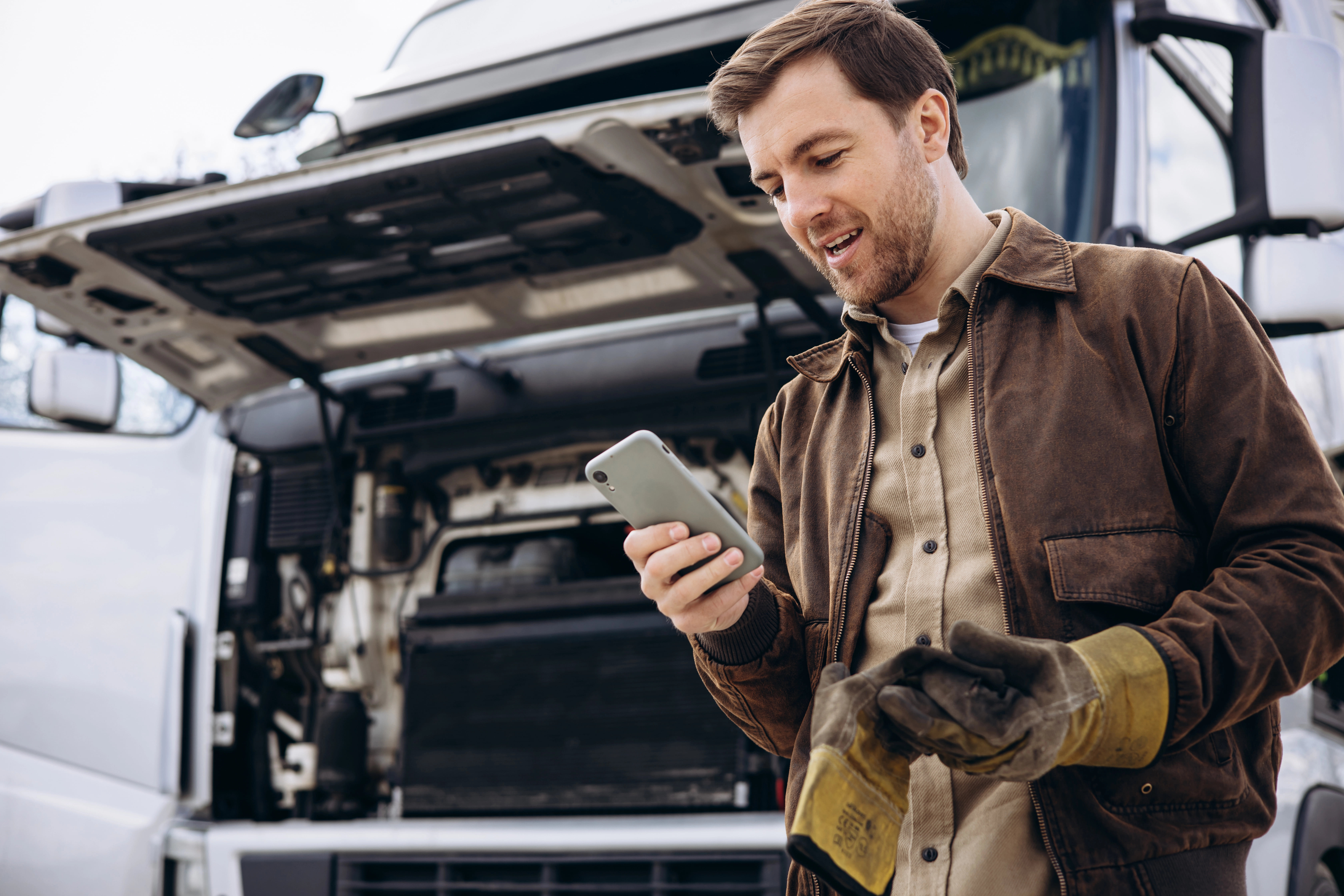 Commercial truck driver with gloves looking at his phone next to a a semi-truck