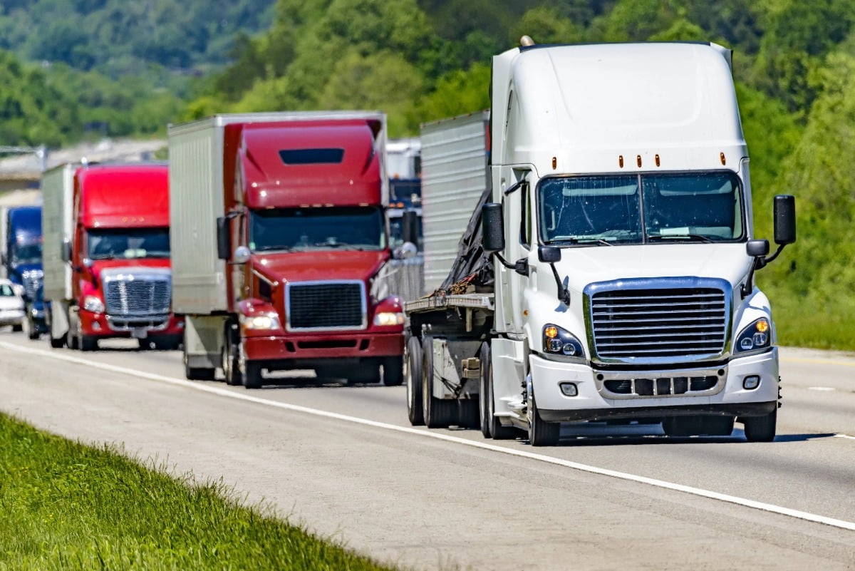 Three Commercial trucks are driving down highway near tree line