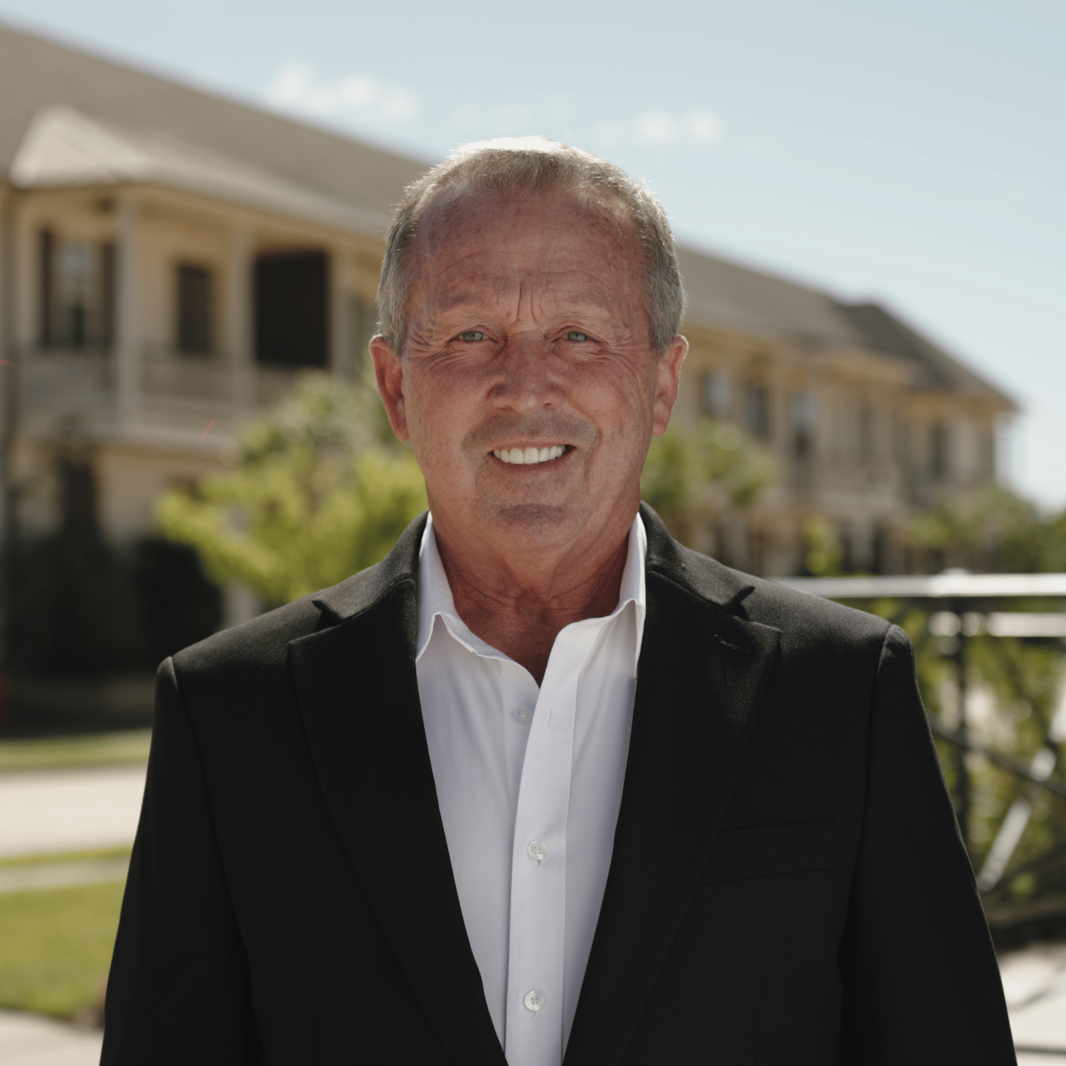Professional business portrait of a real estate broker wearing a black suit outdoors with residential buildings softly blurred behind him