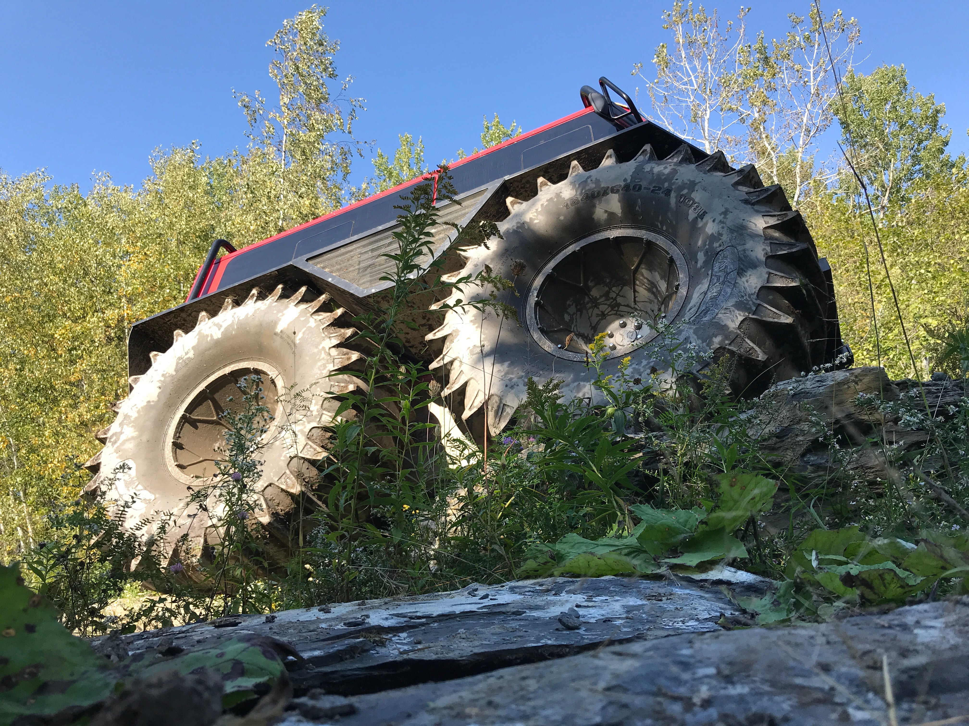 Low-angle view of an amphibious all-terrain vehicle with large lugged flotation tires climbing over rocky ground in front of trees