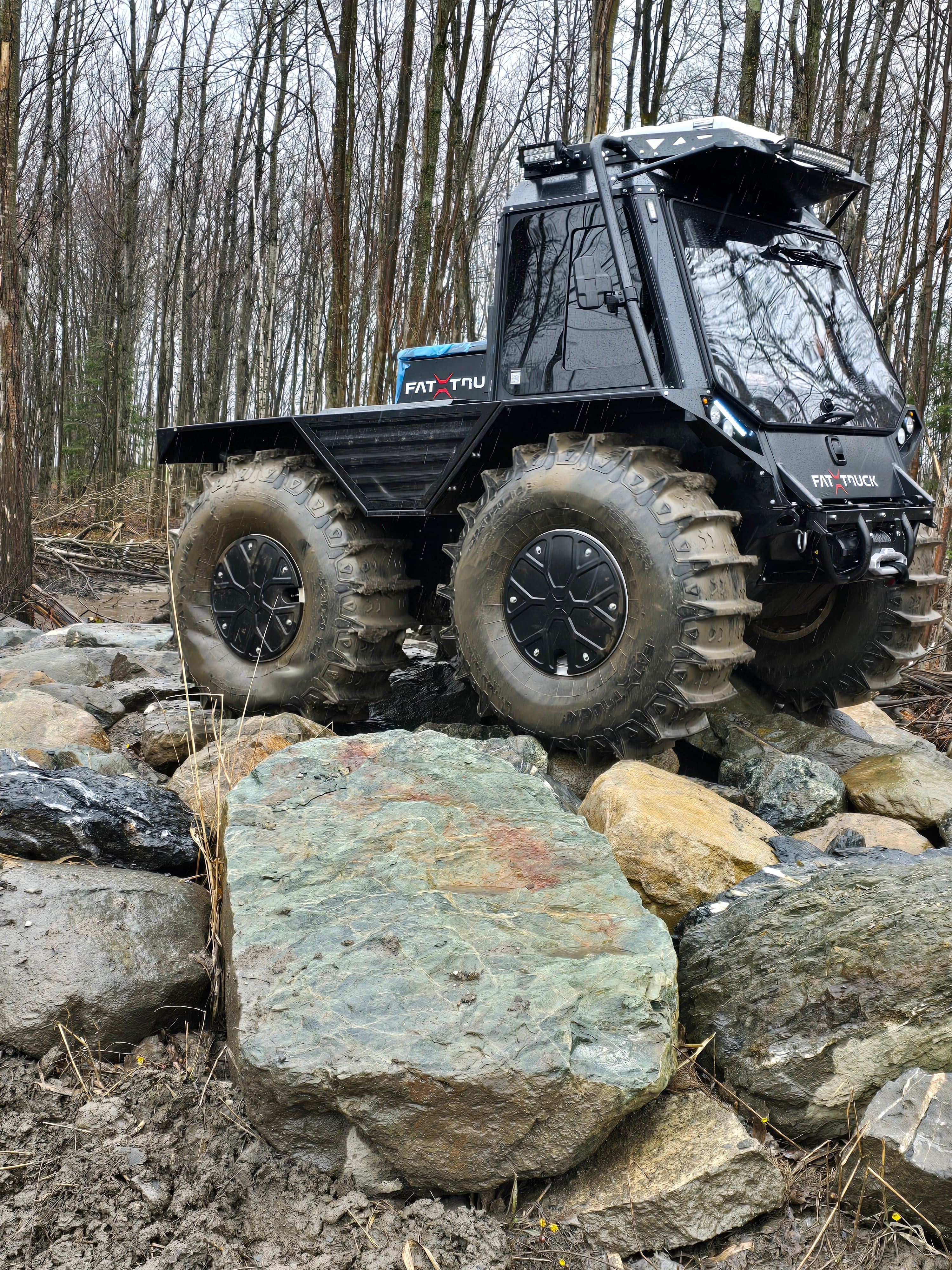 Black Fat Truck amphibious all-terrain vehicle with an enclosed cab and oversized tires, parked on rocky terrain.
