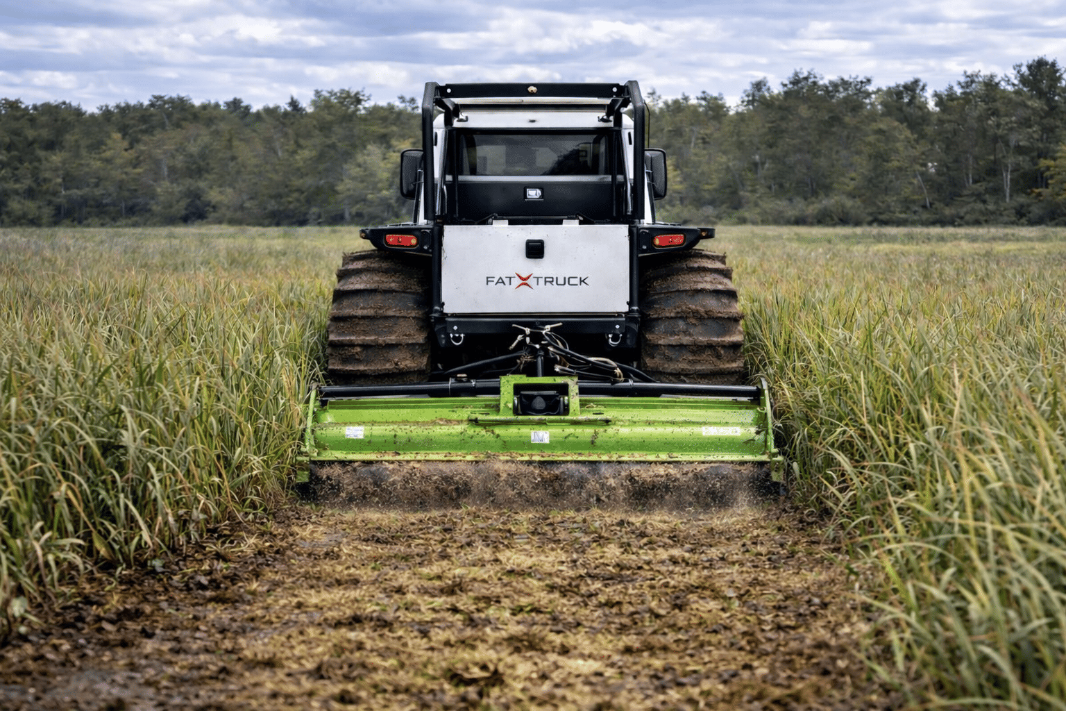 Rear view of a Fat Truck all-terrain vehicle with a mowing attachment, working in a tall grass field beside a tree line