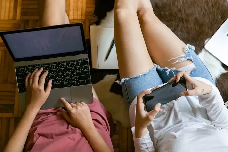 From above of crop girlfriends in home clothes sitting on floor with laptop and smartphone while spending time together and planning weekend