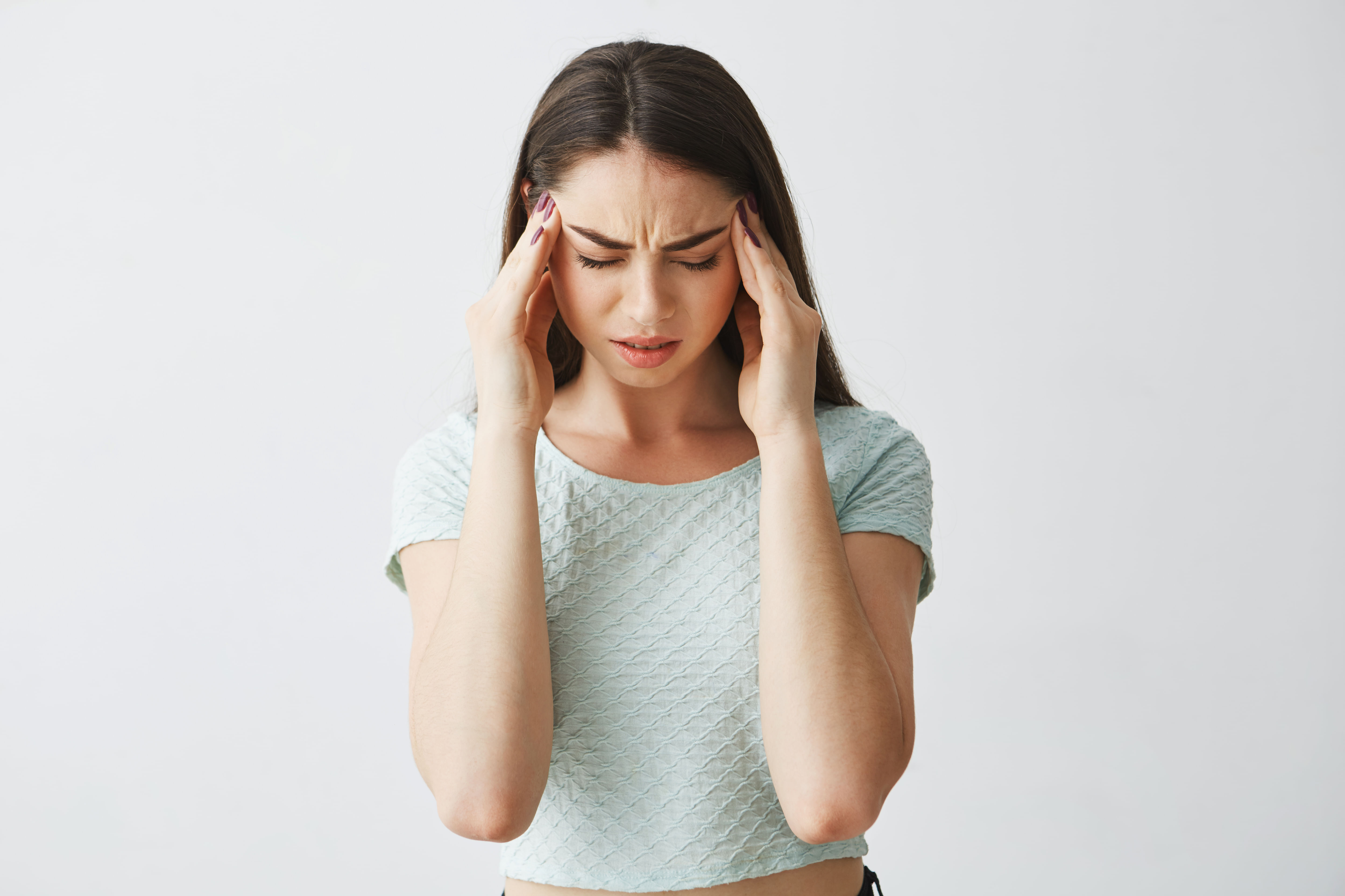 A close-up of a frustrated-looking young woman with dark hair, wearing a light-colored shirt, pressing her fingers against her temples as if she has a severe headache.