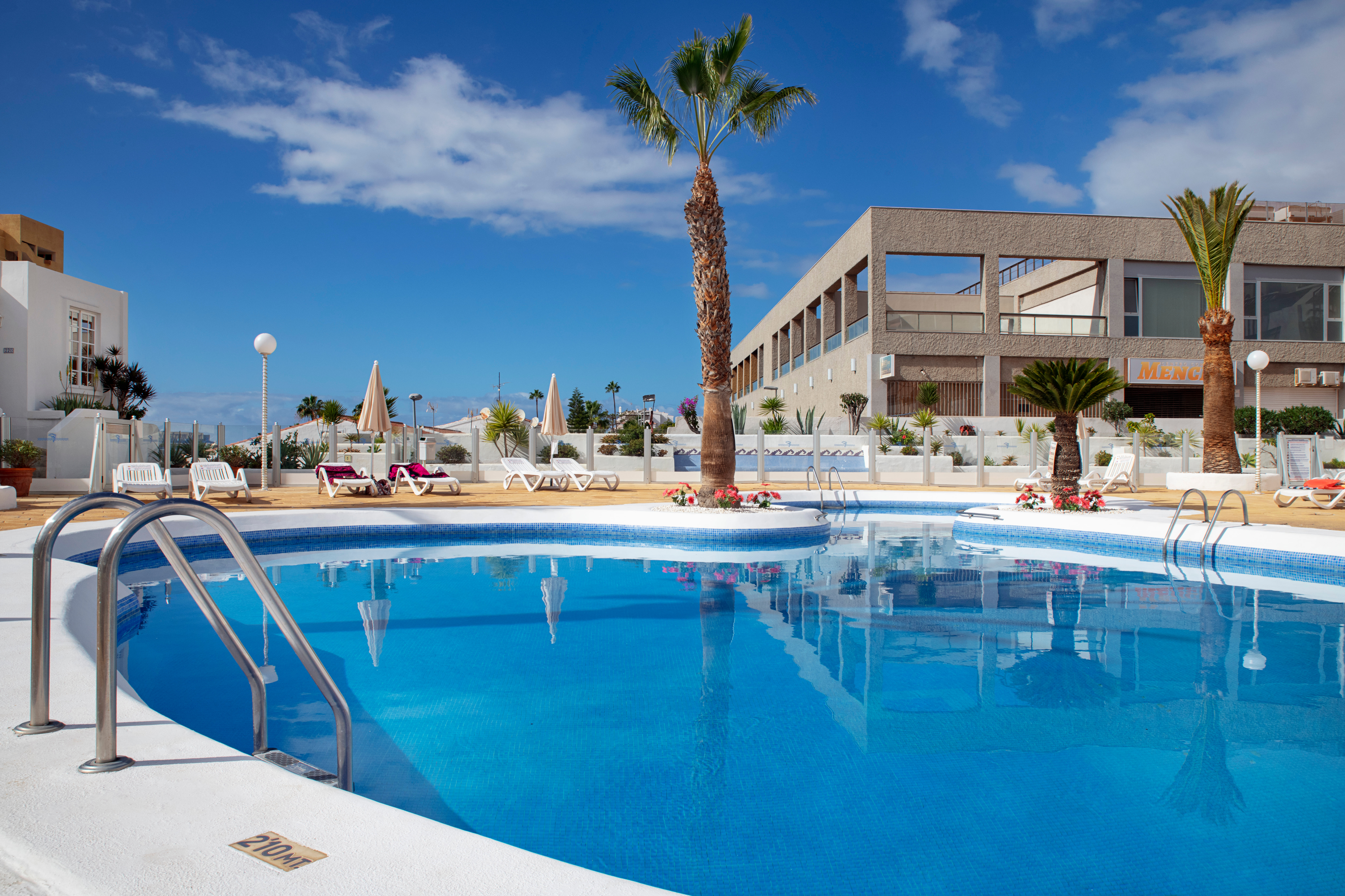Outdoor resort swimming pool surrounded by palm trees, lounge chairs, and modern buildings