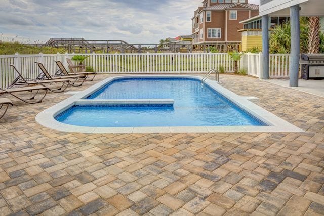 Outdoor swimming pool with tanning ledge, surrounded by a tan paver patio, white fence, lounge chairs, and a coastal home with palm trees in the background