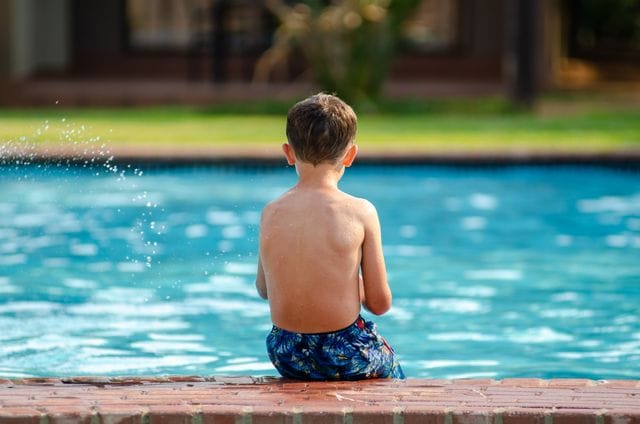 Young boy sitting on the edge of a backyard pool, viewed from behind, with water splashing nearby.