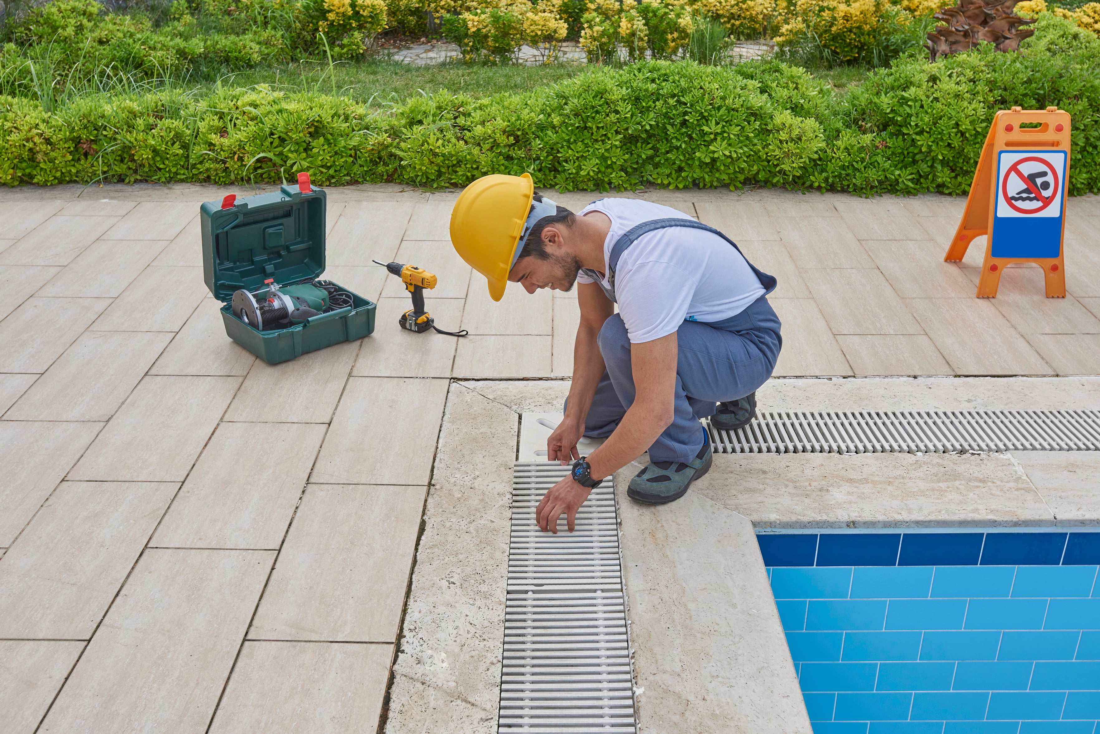 Pool maintenance worker inspecting a drainage grate beside a swimming pool with repair tools nearby