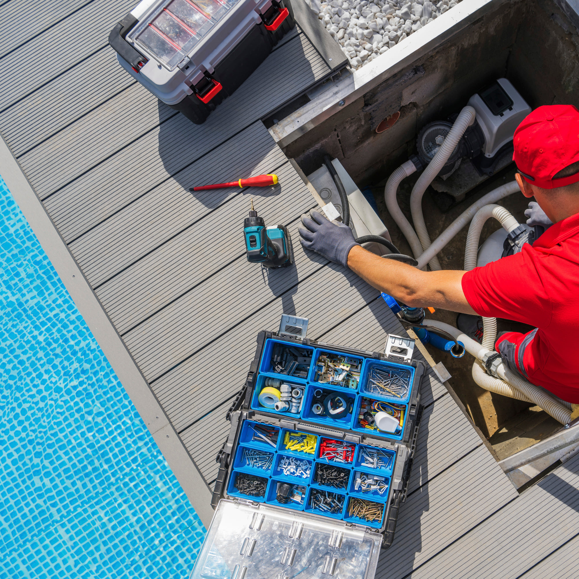Overhead view of a technician repairing pool pump plumbing in an equipment compartment beside the pool, with an open parts organizer
