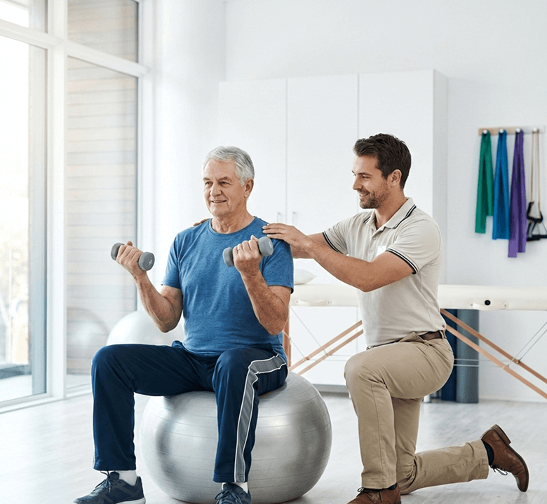 An elderly man sitting on medicinal ball whiling lifting light weights