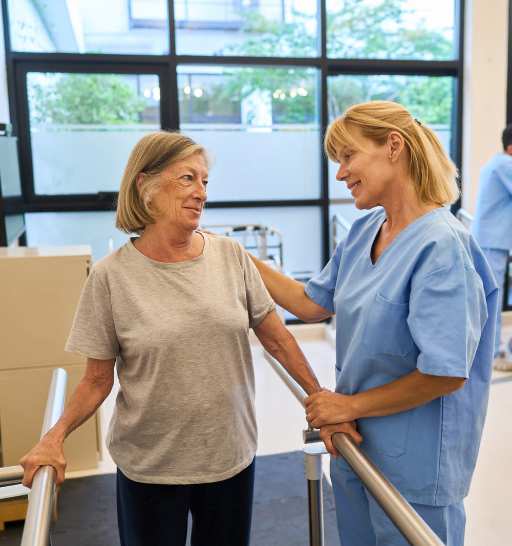 A Blonde haired phyiscal therapist giving care while patient holds onto railings