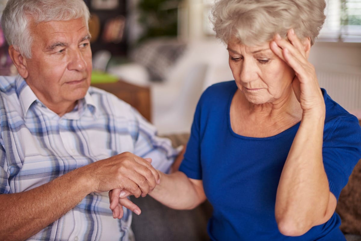 Elderly man watching over elderly woman who is touching her head in pain