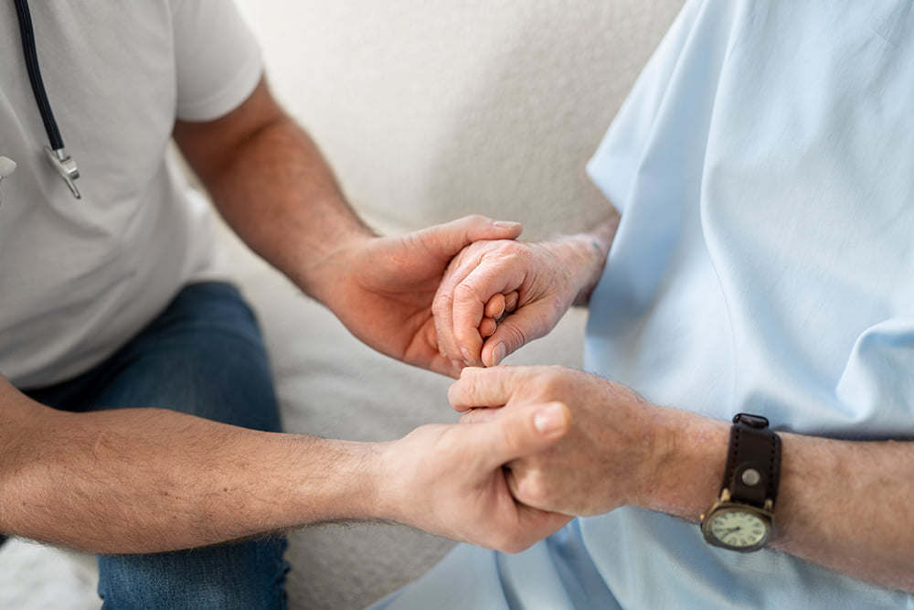 A man is a white t-shirt and blue jeans holding hands with a patient in a hospital gown