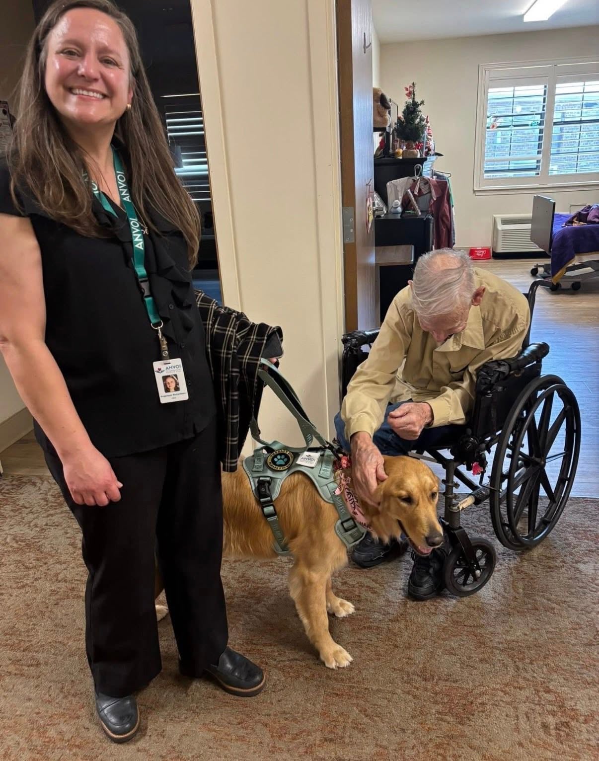 An elderly man sitting in a wheelchair leaning forward to gently pet the head of a golden retriever wearing a therapy dog harness