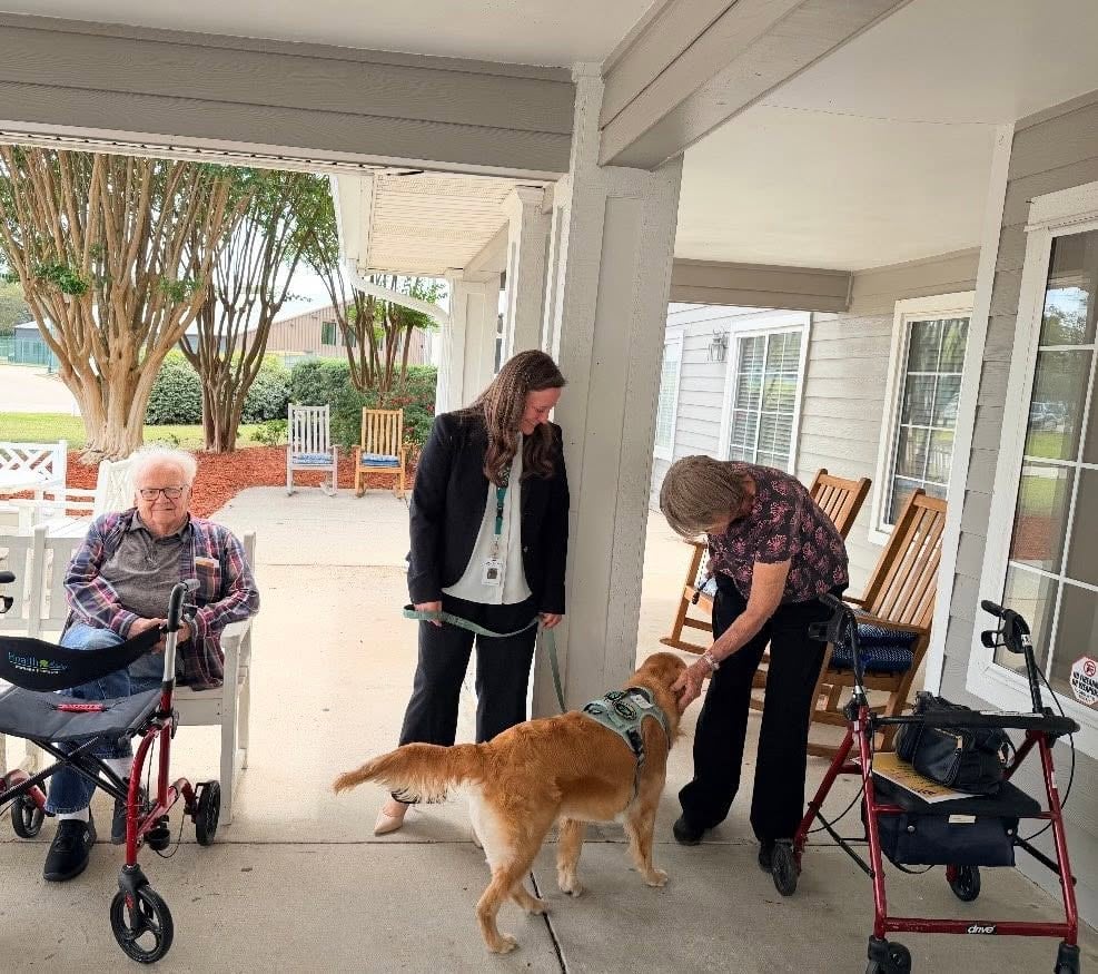 A woman leaning down to pet a golden retriever wearing a harness on a porch outside a facility, while a staff member holds the dog\'s leash and a man with a walker smiles from a nearby chair