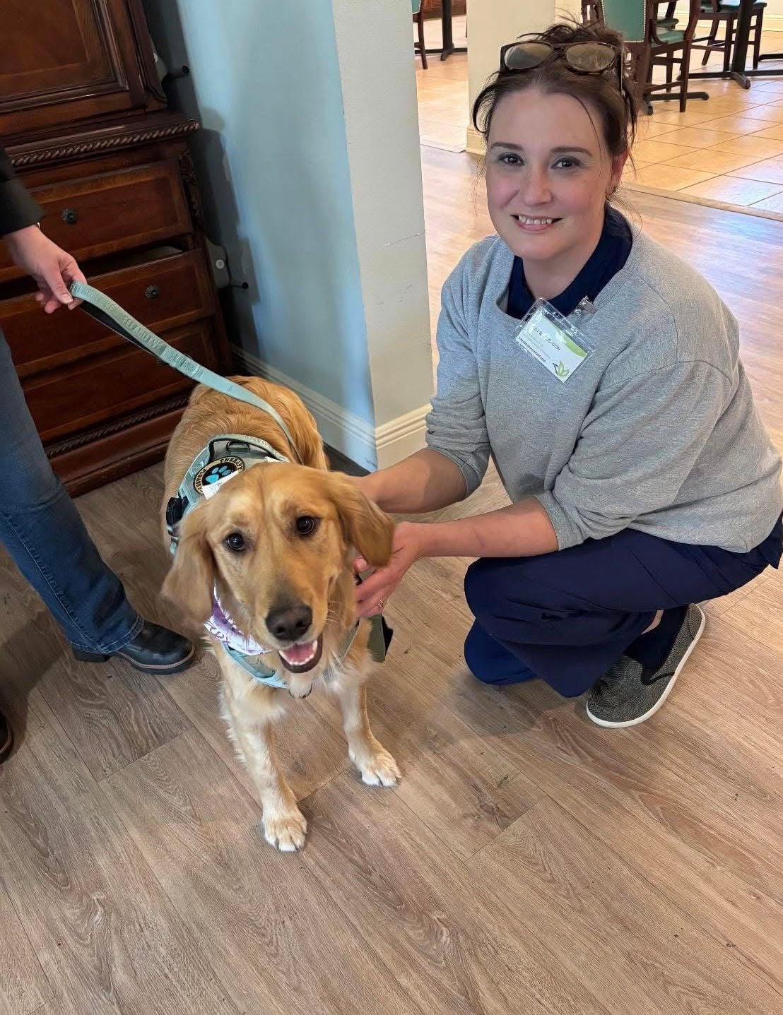 A smiling healthcare staff member kneeling on the floor next to a happy golden retriever wearing a therapy dog harness.