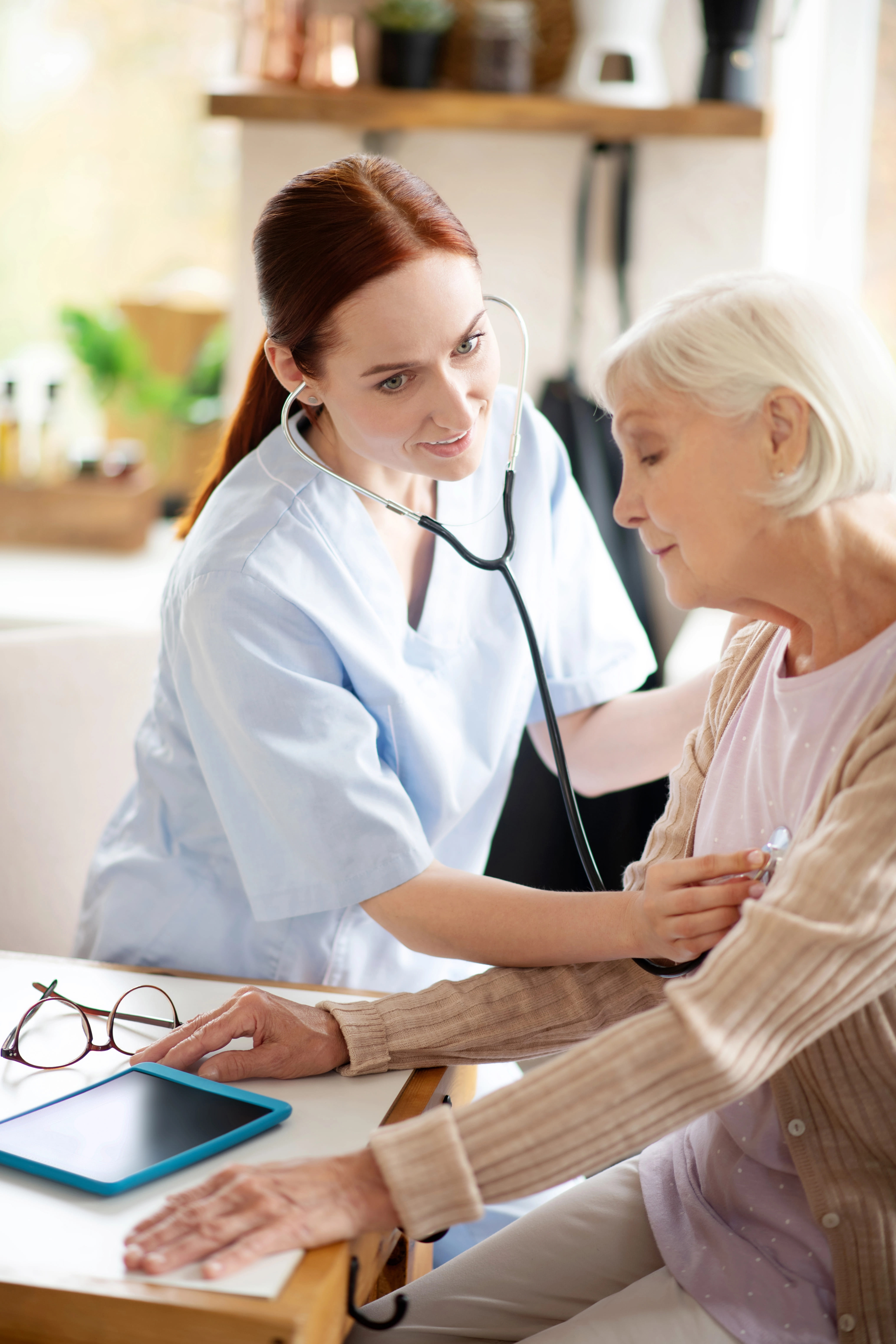 Red-headed hospice care doctor checking the heartbeat of an elderly patient with a stethoscope