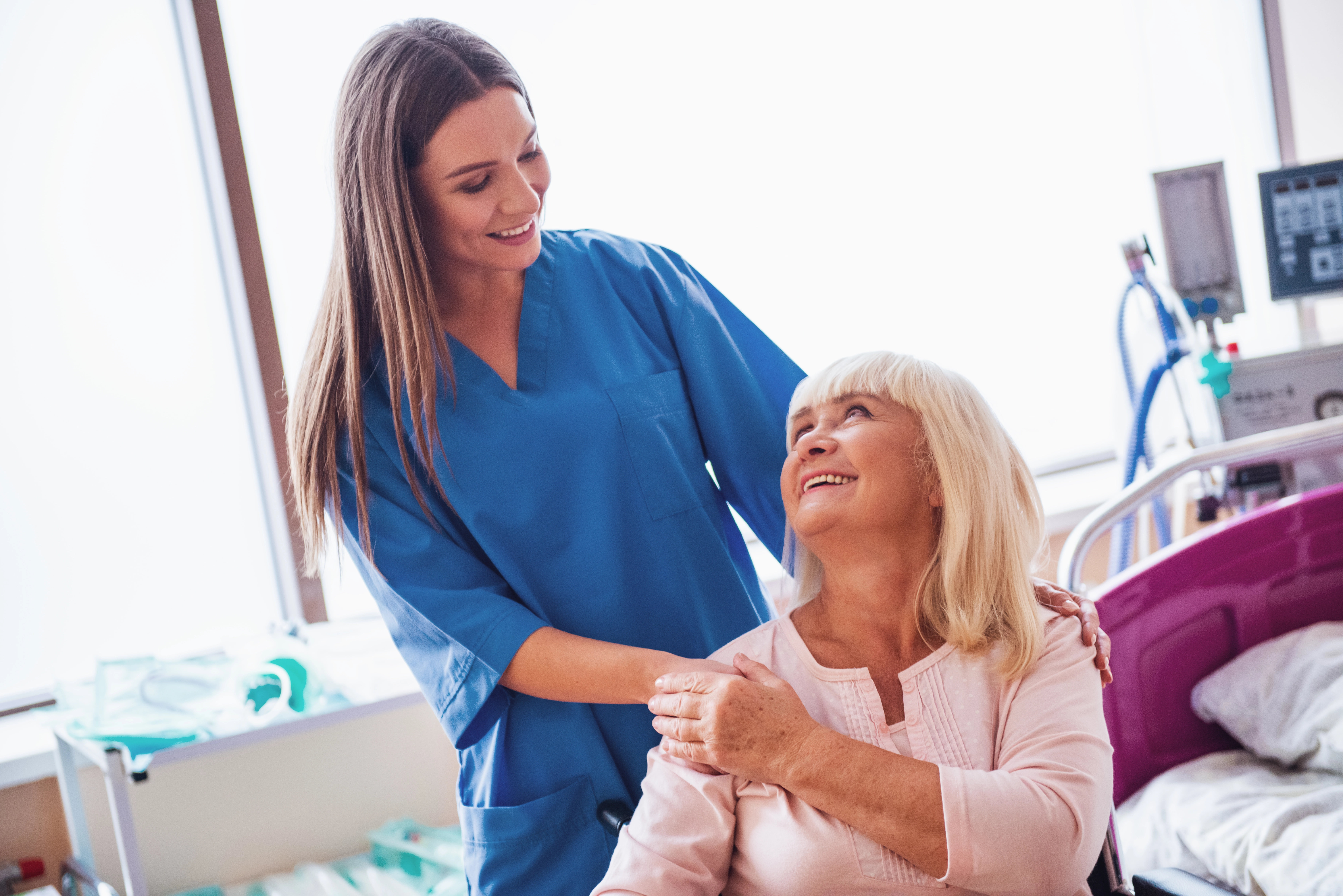 A healthcare professional in blue is holding an elderly woman\'s hand while speaking with her as she sits in a wheelchair