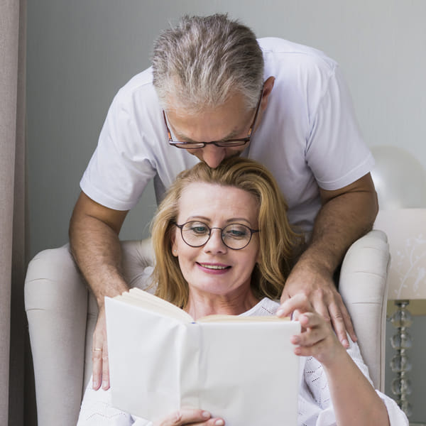 Husband leaning behind his wife to kiss her forehead as she reads a novel