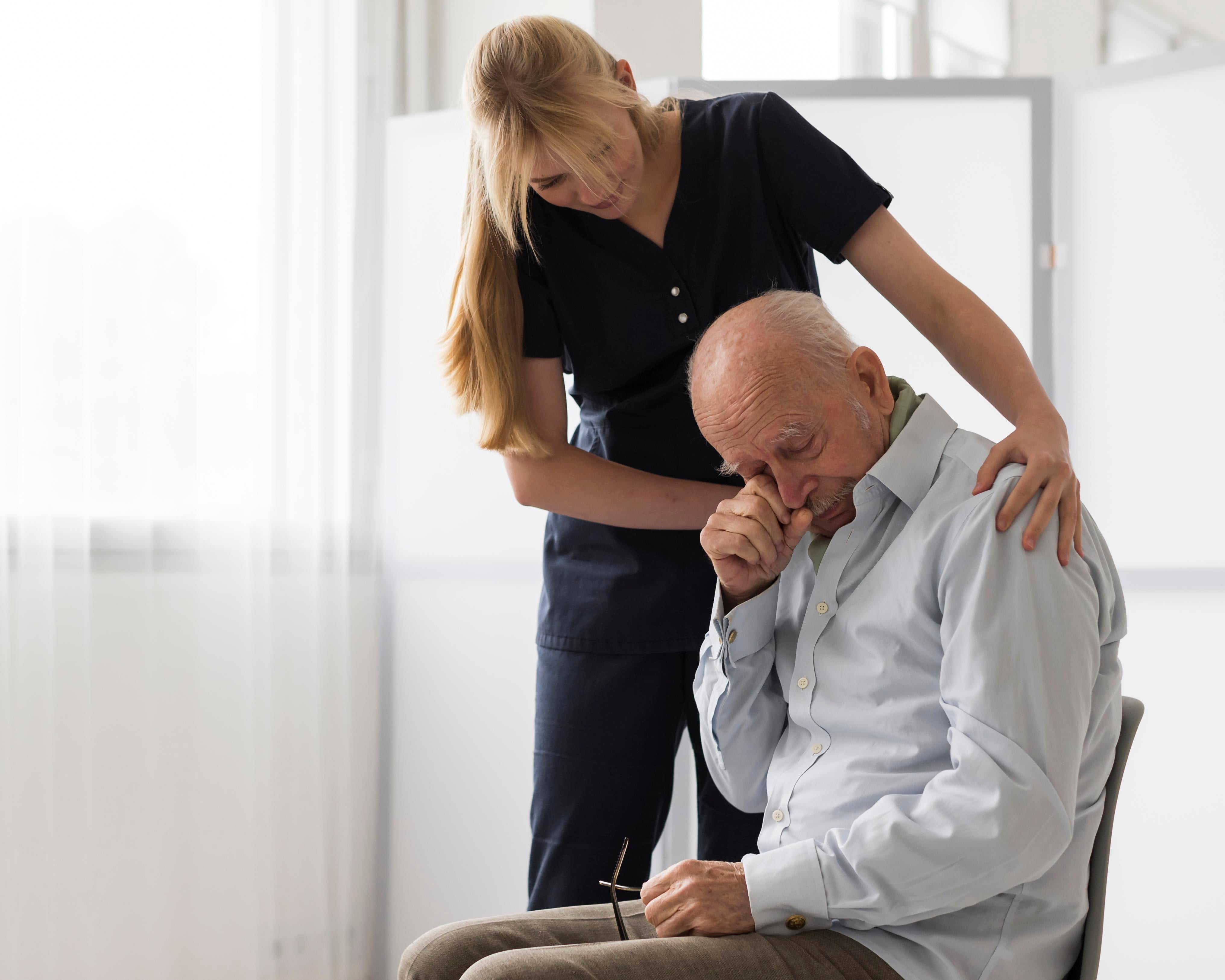 Nurse placing a reassuring hand on an elderly man\'s shoulder as he wipes tears from his eyes