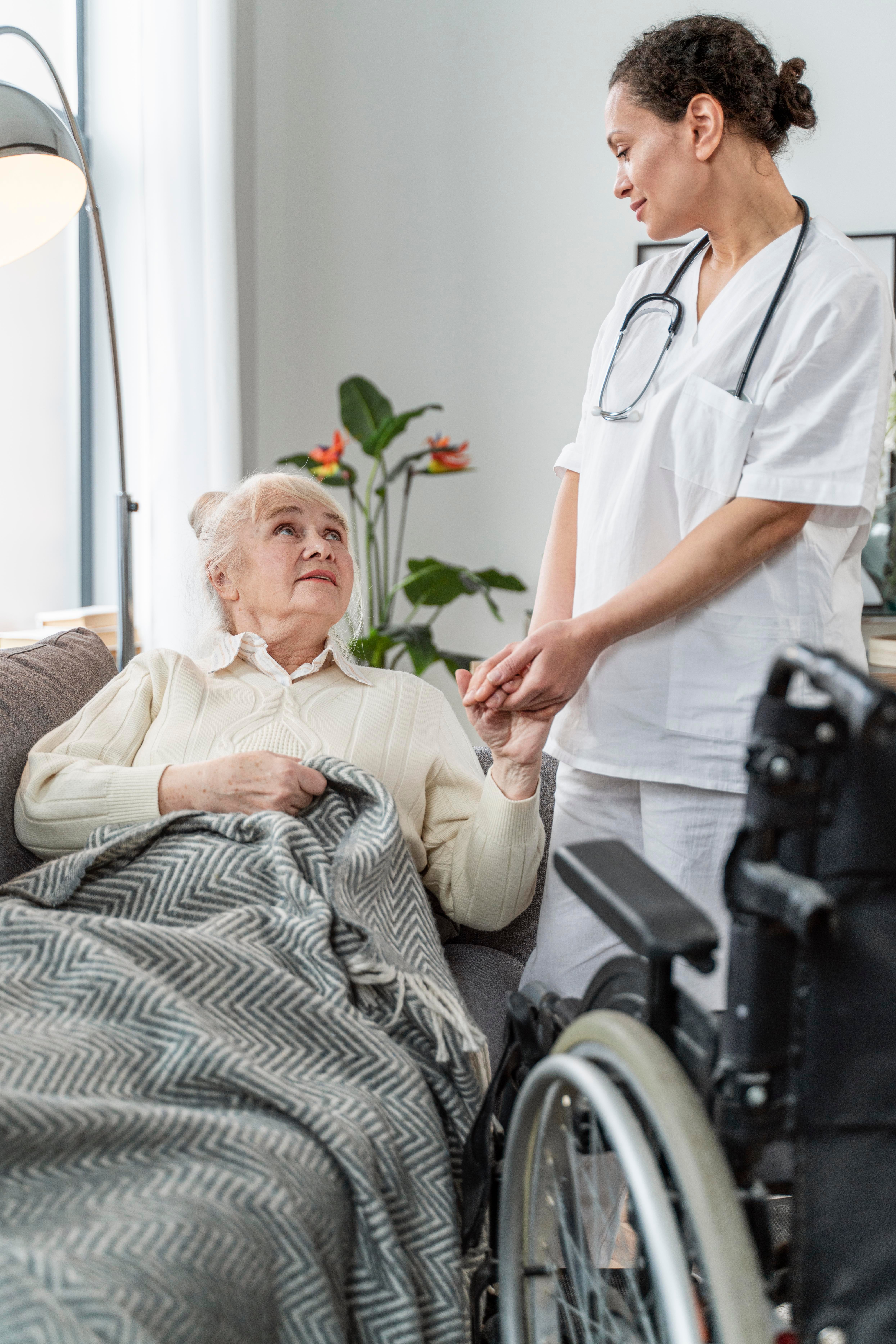 Healthcare professional in white scrubs holding an elderly woman\'s hand while speaking with her beside a wheelchaice