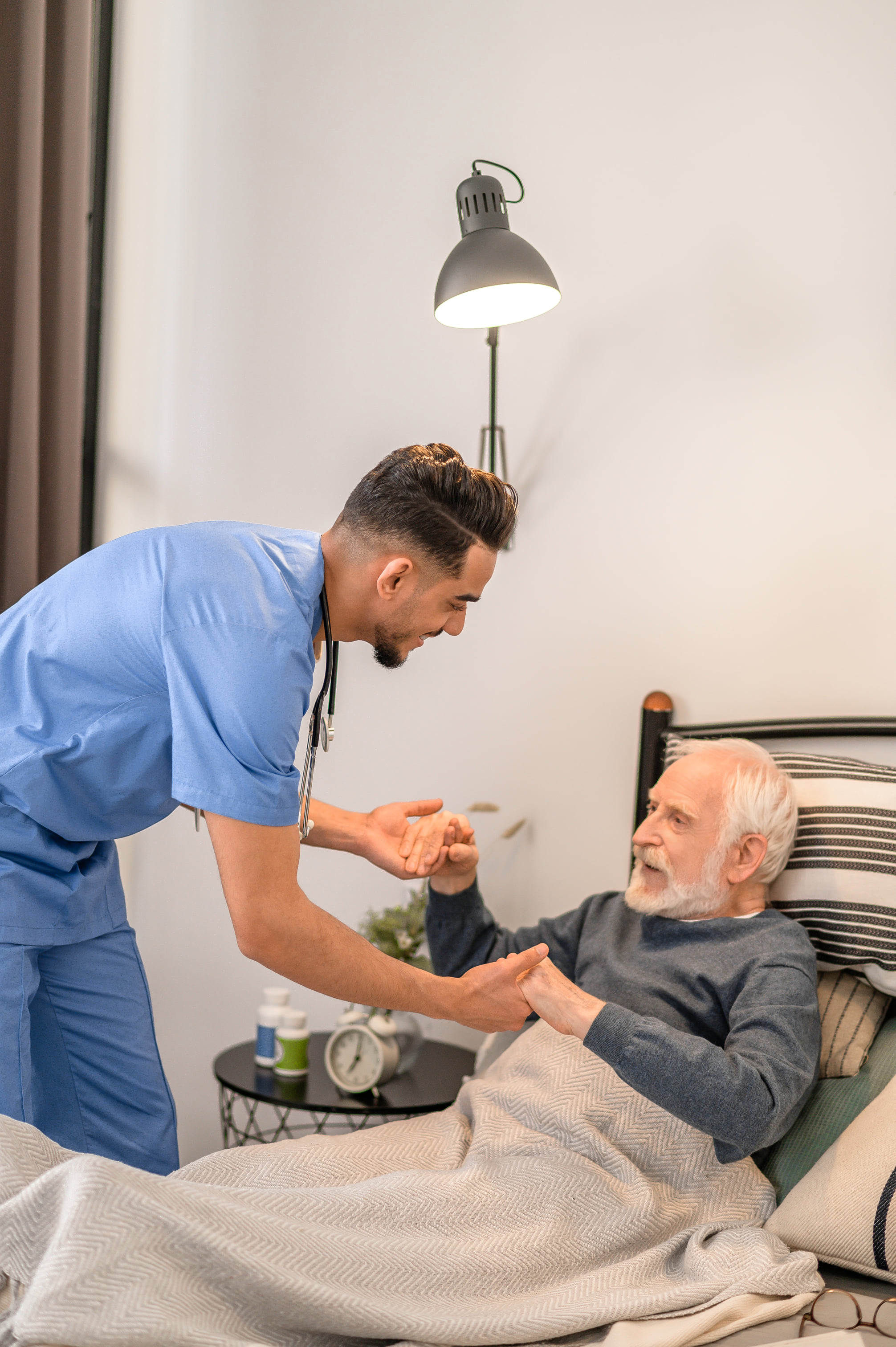 Home healthcare nurse leaning over to help an elderly man sit up in bed while holding his hands