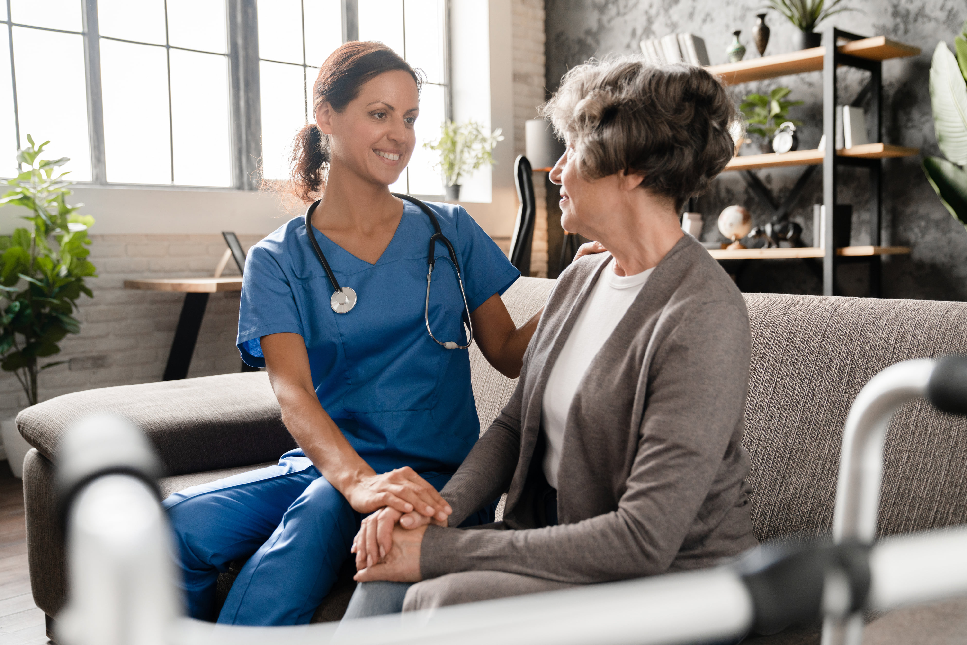 Home healthcare nurse sitting beside an elderly woman on a sofa and holding her hands during a caring conversation