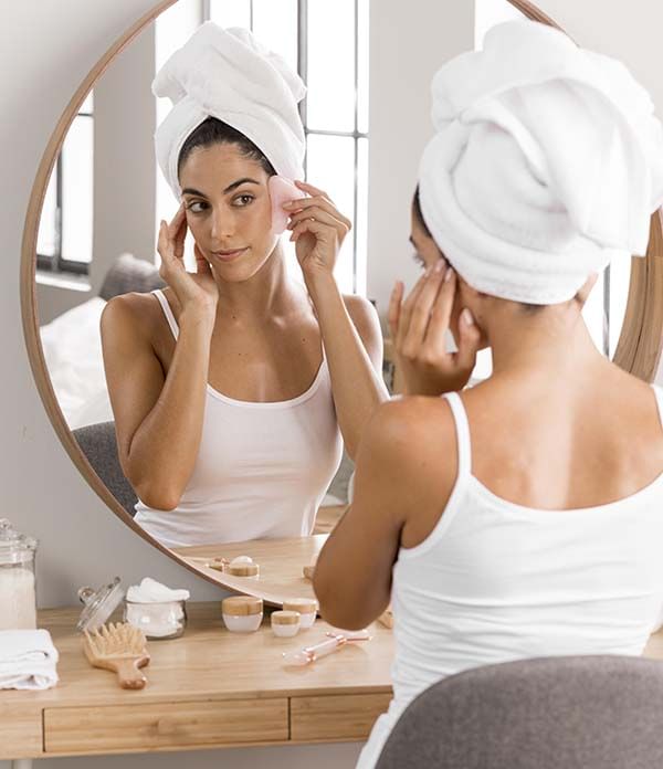 Woman with towel-wrapped hair applying skincare product while looking in bathroom mirror. Woman with towel-wrapped hair applying skincare product while looking in bathroom mirror.