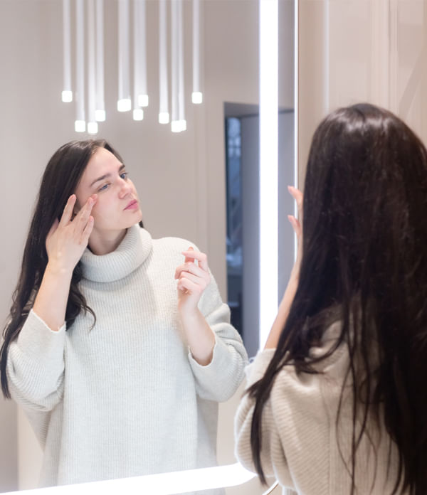 Woman in cozy sweater applying cream under her eyes while standing in front of a mirror. Woman in cozy sweater applying cream under her eyes while standing in front of a mirror.