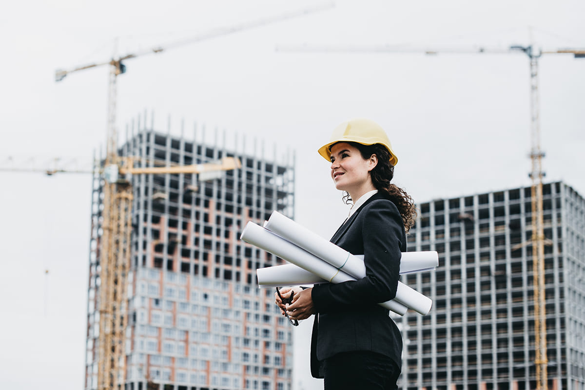 Professional archtiect holding blueprints as she looks over steel-framed high-rises during the frame building process